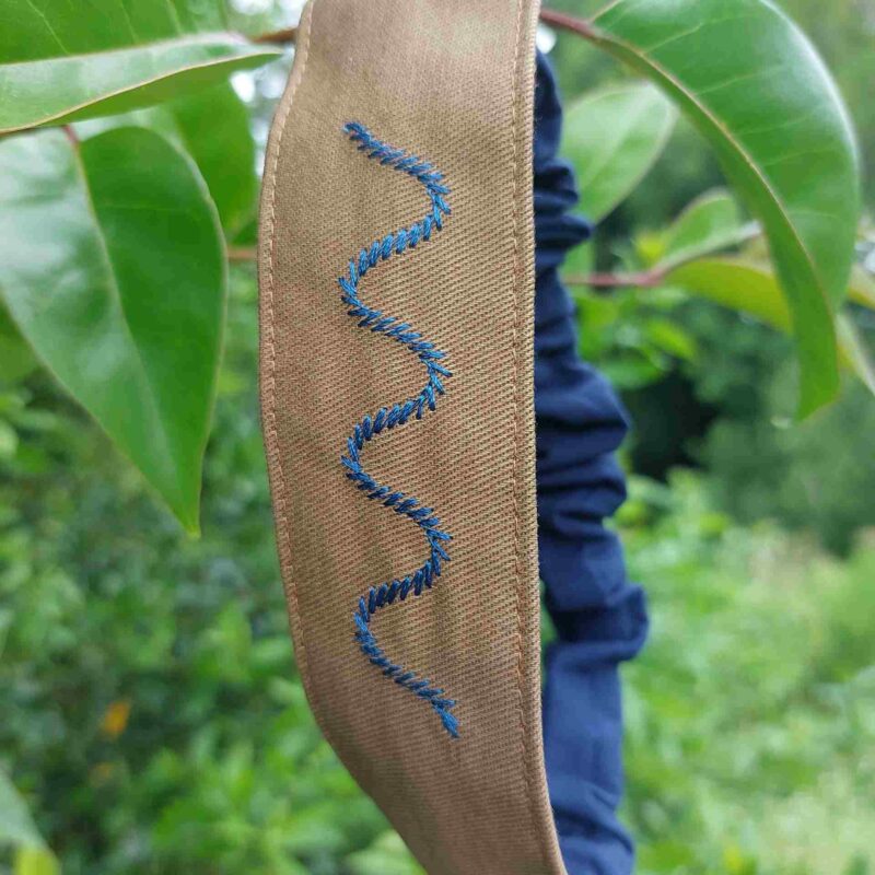 Bandeau Mer Serpent cousu en coton à Bayonne sur un tissu marron. Il est brodé avec un motif en bleu représentant les vagues de la mer à la façon de l'ondulation d'un serpent du fait de l'utilisation de son point de broderie.